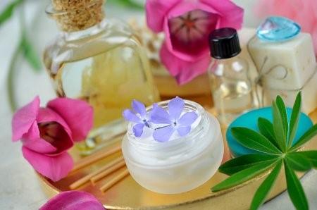Na zdjęciu widoczna jest kompozycja z różnych produktów do pielęgnacji, ułożona na złotej, okrągłej tacy. W centrum znajduje się otwarty, mały słoiczek z białym kremem, na którym leżą dwa małe, fioletowe kwiatki. Dookoła niego rozmieszczone są inne produkty: buteleczki z olejkami, kawałki mydła, a także ozdobne, różowe kwiaty. Całość sprawia wrażenie naturalnych, delikatnych i luksusowych kosmetyków do pielęgnacji skóry.